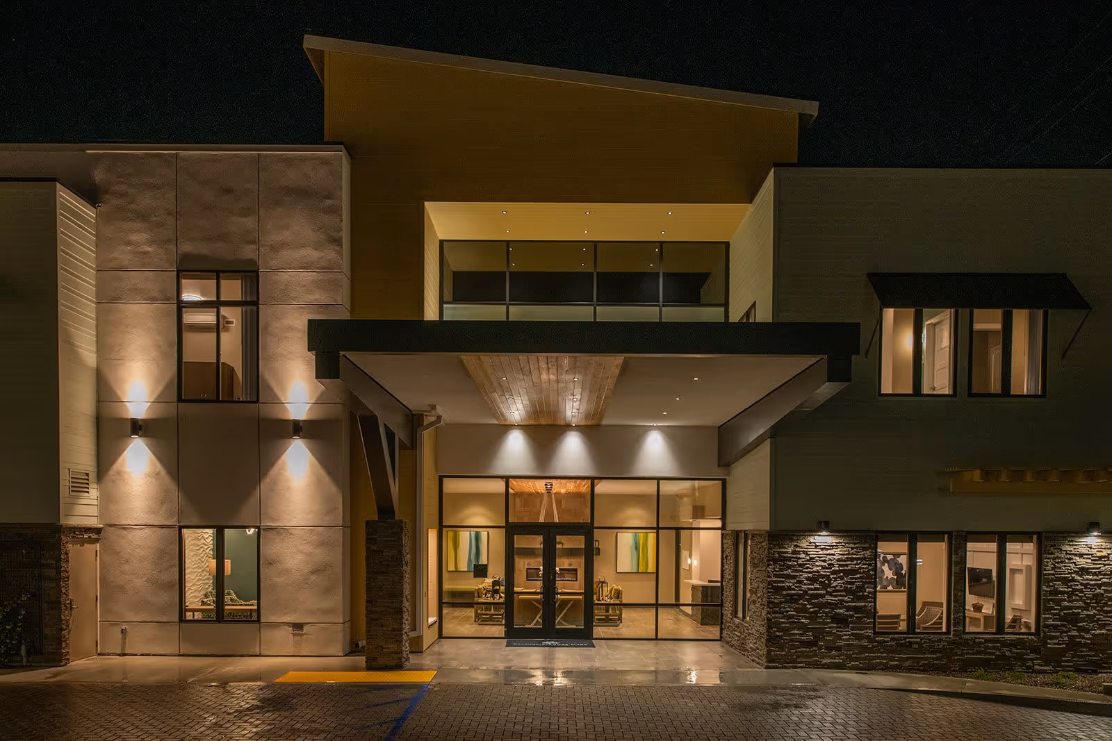 Night view of the entrance to a modern building with large glass doors and windows, exterior lights illuminating the facade, and a covered entryway with wooden ceiling panels.