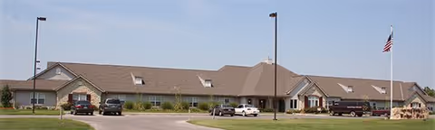 Single-story brick-and-stone senior living building with parked cars, a flagpole, and a front driveway under a clear sky.