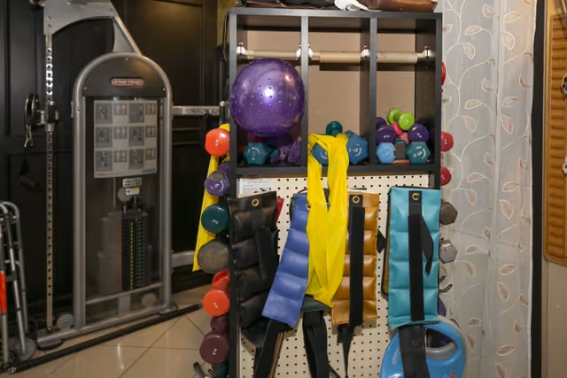 A fitness equipment rack holding various dumbbells, resistance bands, weighted ankle straps, and a purple exercise ball in a gym area with exercise machines in the background.