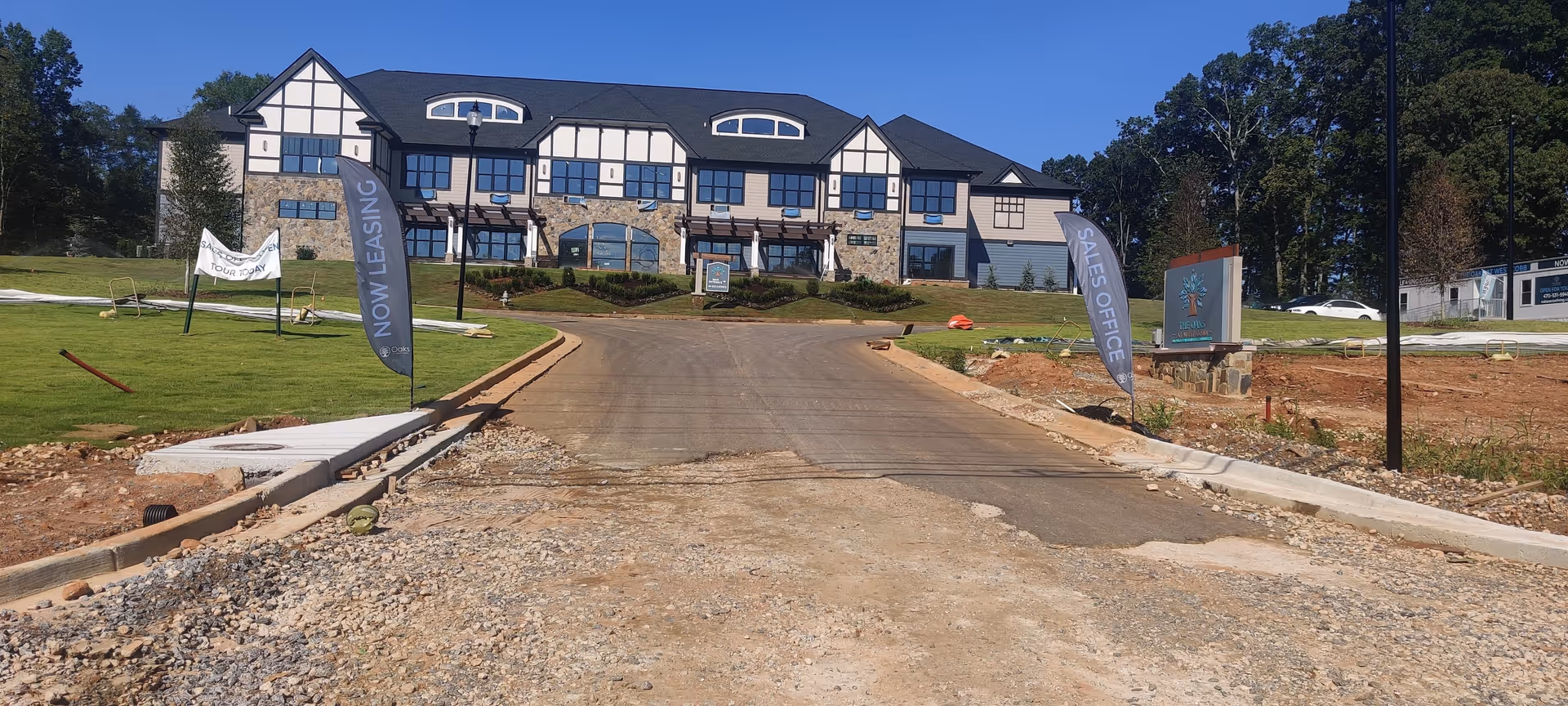 View of a large two-story building with a stone and beige exterior at the end of a paved driveway under construction. There are two vertical flags on either side of the driveway, one reading 'NOW LEASING' and the other 'SALES OFFICE'. A sign near the right flag displays the Oaks at West Cobb logo. The surrounding area has patches of grass and some construction materials.