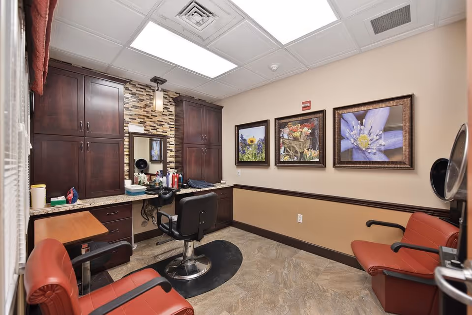 Interior view of a small salon or grooming room with a black salon chair in front of a mirror and countertop with hair products. The room has dark wooden cabinets, beige walls with three framed floral pictures, and two red chairs for waiting or clients. The floor is tiled and the ceiling has fluorescent lighting.