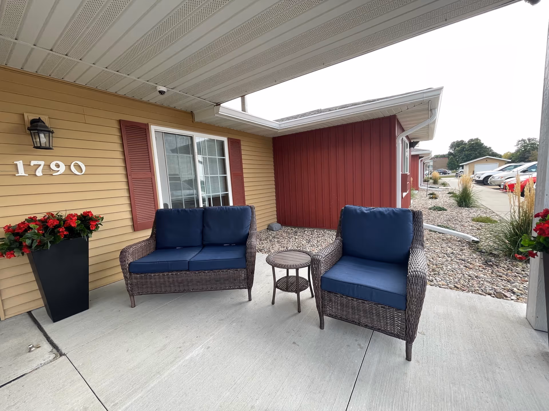 Covered outdoor seating area with a wicker loveseat and chair with blue cushions, a small round table between them, a beige building wall with the number 1790 and red shutters on the window, a black planter with red flowers, and a gravel landscaping area with plants and parked cars in the background.