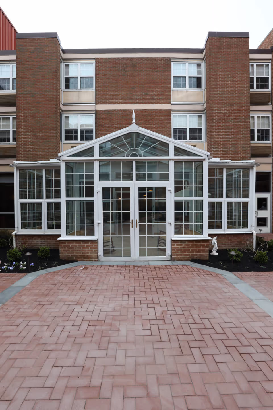 Exterior view of a brick building with a glass entrance structure featuring double doors and multiple windows. The entrance is surrounded by a paved walkway and small landscaped areas with plants.