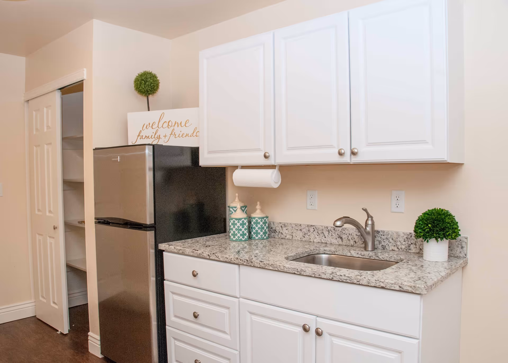 A small kitchen area with white cabinets, a granite countertop, a stainless steel sink with a faucet, and a stainless steel refrigerator. There are decorative items on the countertop including two patterned canisters and a small green plant. A sign above the refrigerator reads 'welcome family + friends'. There is also a pantry with sliding doors next to the refrigerator.