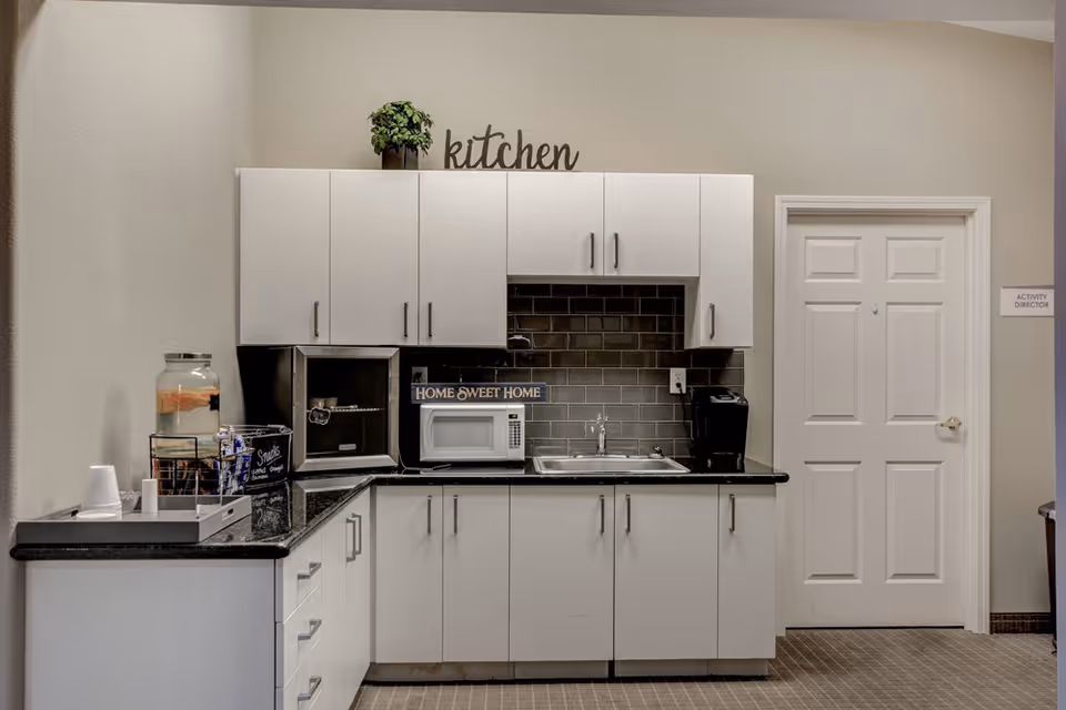 A small kitchen area with white cabinets, a black countertop, a microwave, a sink, a coffee maker, and a water dispenser with cups. There is a decorative sign that says 'HOME SWEET HOME' and another sign above the cabinets that says 'kitchen'. A door labeled 'ACTIVITY DIRECTOR' is visible on the right side.