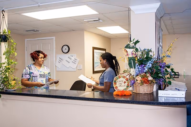 Two women standing and talking across a reception desk in a well-lit room. One woman is wearing a patterned scrub top and holding papers, while the other woman is in a solid-colored scrub top also holding papers. The reception desk has a large floral arrangement and a small decorative sign that says 'Give Thanks'. There are plants hanging and placed around the area, a clock on the wall, and some papers pinned to a bulletin board behind the desk.