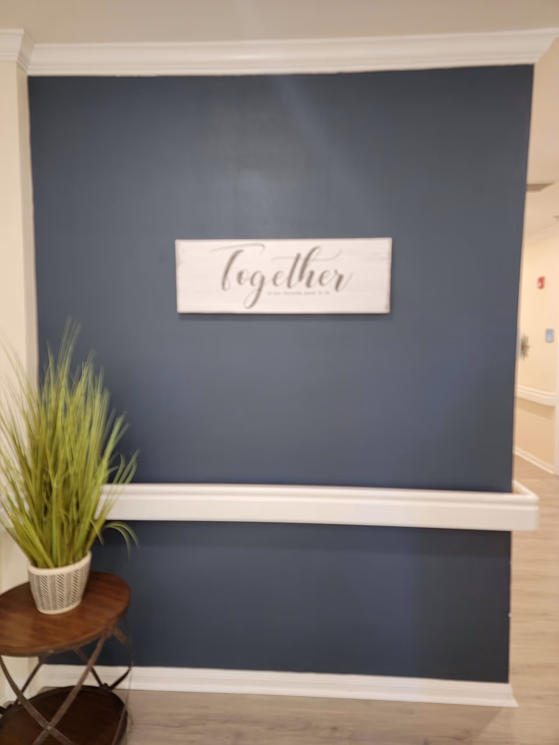 A hallway with a dark blue accent wall featuring a white wooden sign that says 'Together'. There is a small wooden table with a potted green plant on the left side of the wall. The floor is light-colored wood, and the hallway extends to the right with light-colored walls.