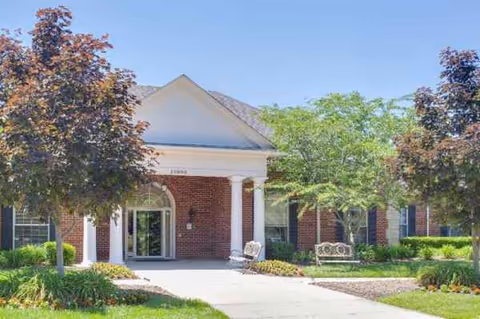 Brick building entrance with white columns, walkway, benches, and trimmed trees on a sunny day.