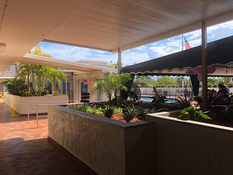 Covered outdoor walkway with planter boxes filled with green plants and flowers. In the background, there is a swimming pool area with a black canopy and people sitting underneath it. The sky is partly cloudy with an American flag visible in the distance.