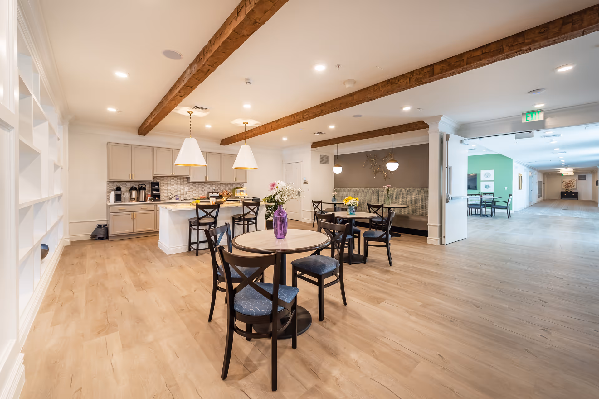 A spacious and well-lit common area in a senior living facility featuring wooden floors, exposed wooden ceiling beams, and multiple round tables with chairs. There is a kitchen area with cabinets, a countertop, and pendant lights. The room opens into a hallway with additional seating and green accent walls.