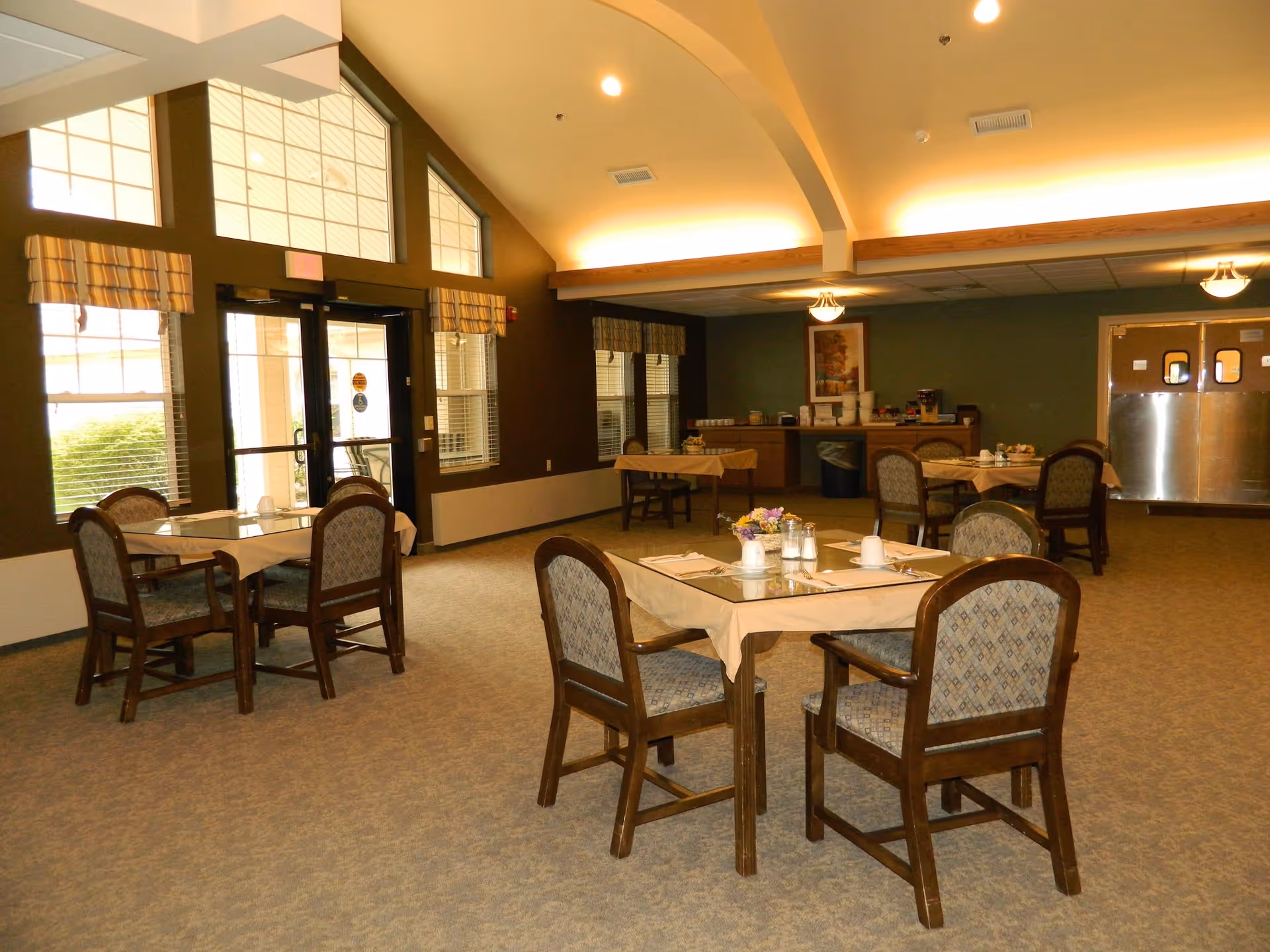 Dining area with several small tables and chairs beneath a vaulted ceiling and large windows.