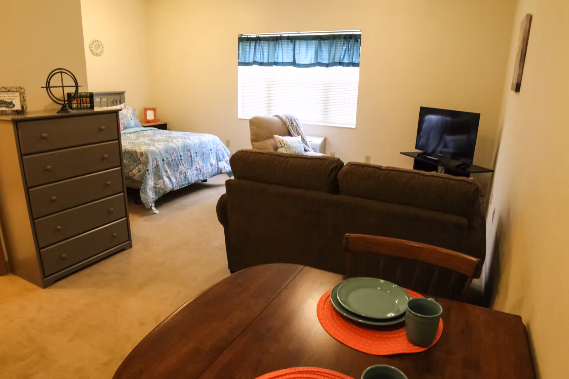 A cozy studio apartment with a bed covered in a blue patterned quilt, a brown couch facing a flat-screen TV on a glass stand, a beige armchair with a decorative pillow, a wooden dresser, and a small dining table set with green plates and mugs on red placemats. A window with blue curtains lets in natural light.
