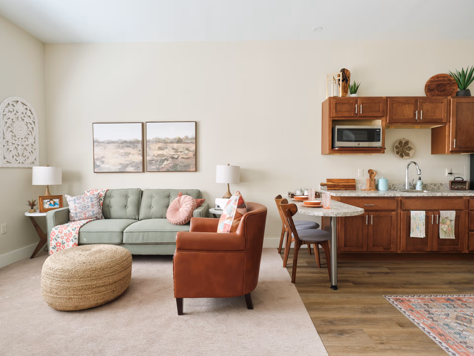 A cozy living area with a light green tufted sofa adorned with patterned pillows and a pink round pillow, a brown leather armchair with a colorful pillow, a woven ottoman, and two table lamps on side tables. The room features light beige walls with two landscape paintings and decorative wall art. Adjacent to the living area is a kitchen with wooden cabinets, a microwave, a sink, and a small dining counter with two chairs set with plates and glasses.