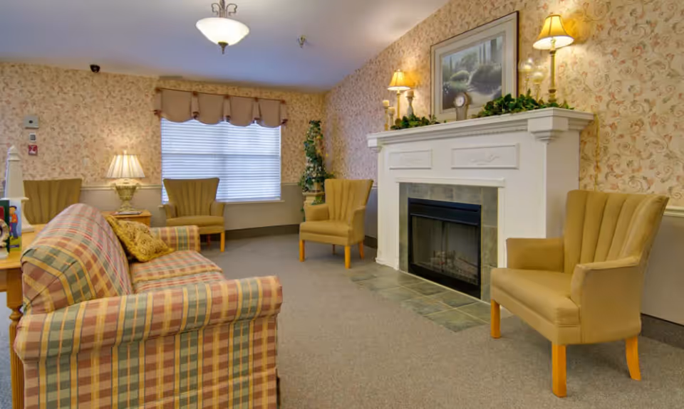 A cozy living room with a plaid sofa, three beige armchairs, a white fireplace with decorative greenery and lamps on the mantel, floral wallpaper, a window with blinds and a valance, and a ceiling light fixture.