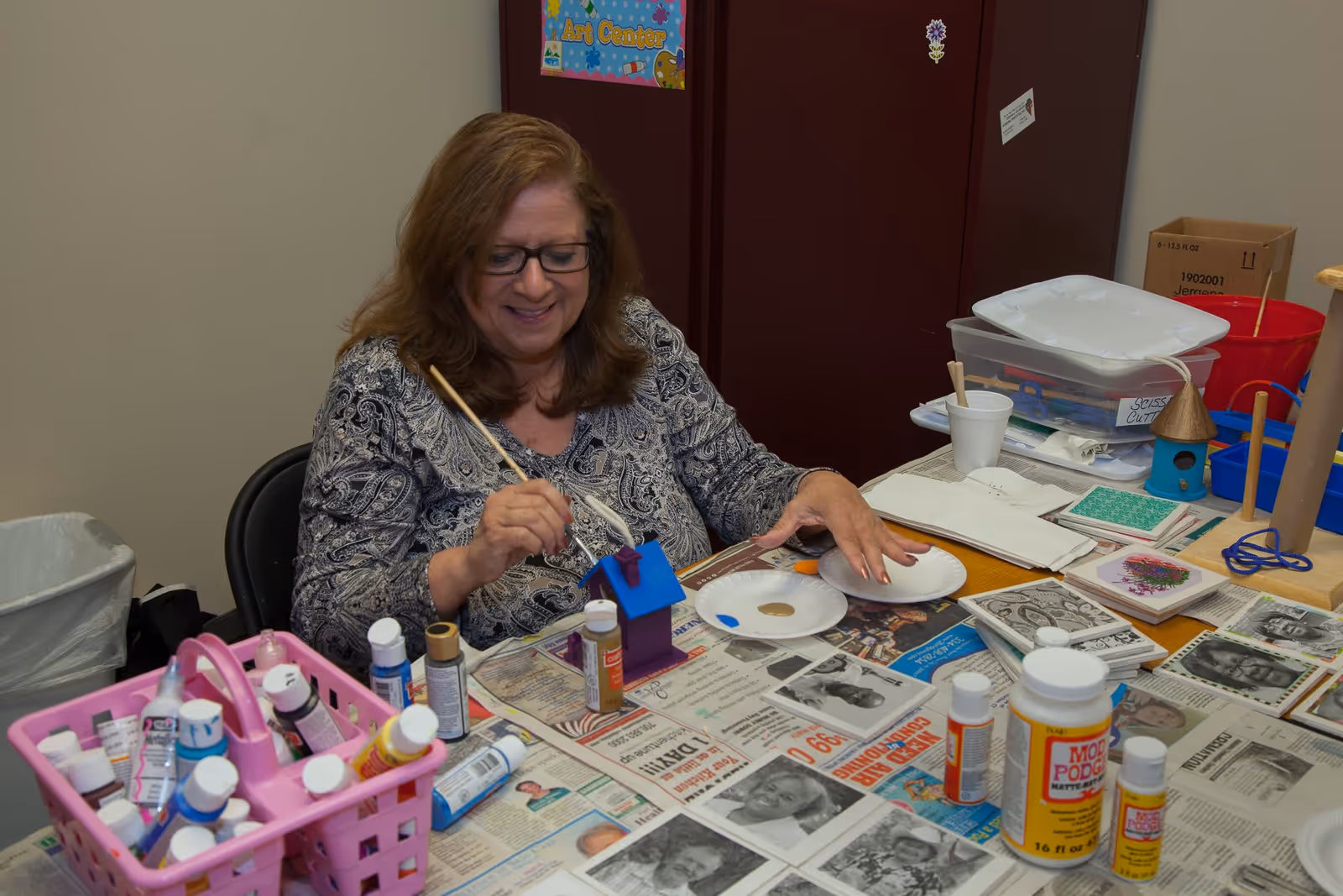 A woman with glasses sitting at a table covered with newspapers and various art supplies, painting a small wooden house blue. The table has bottles of paint, brushes, and other craft materials. The background shows a maroon cabinet and some storage containers.