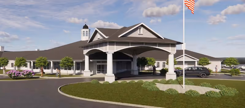 Exterior view of a single-story senior living facility building with a covered entrance, a circular driveway, landscaped greenery, and an American flag on a flagpole under a partly cloudy sky.