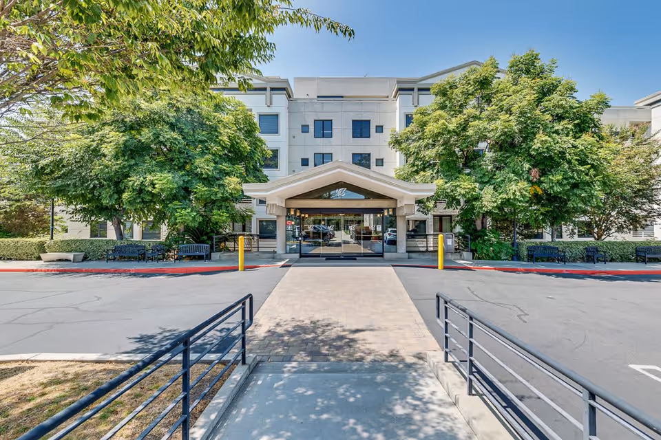 Front exterior view of Sakura Intermediate Care Facility showing a multi-story building with a covered entrance, surrounded by green trees and benches along the sidewalk, under a clear blue sky.