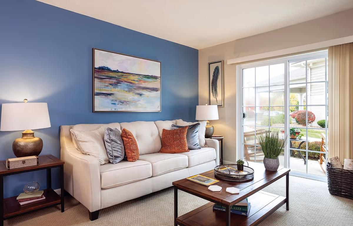 Bright living room with a beige sofa, colorful throw pillows, a wooden coffee table, lamps, artwork on a blue accent wall, and sliding glass doors to a patio.