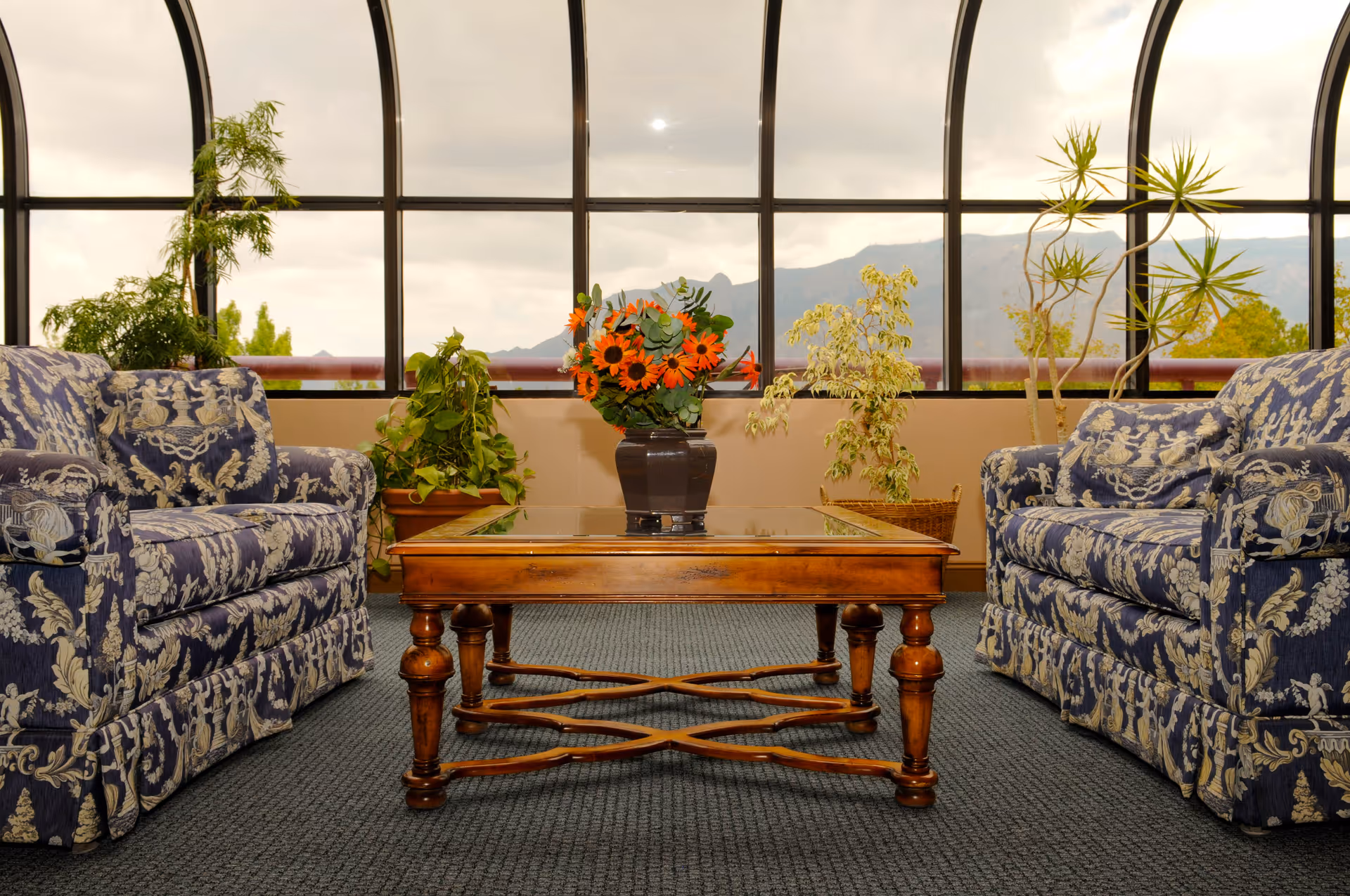 A sunlit sitting area with two patterned armchairs facing a wooden coffee table topped with a vase of orange flowers in front of large arched windows and potted plants.