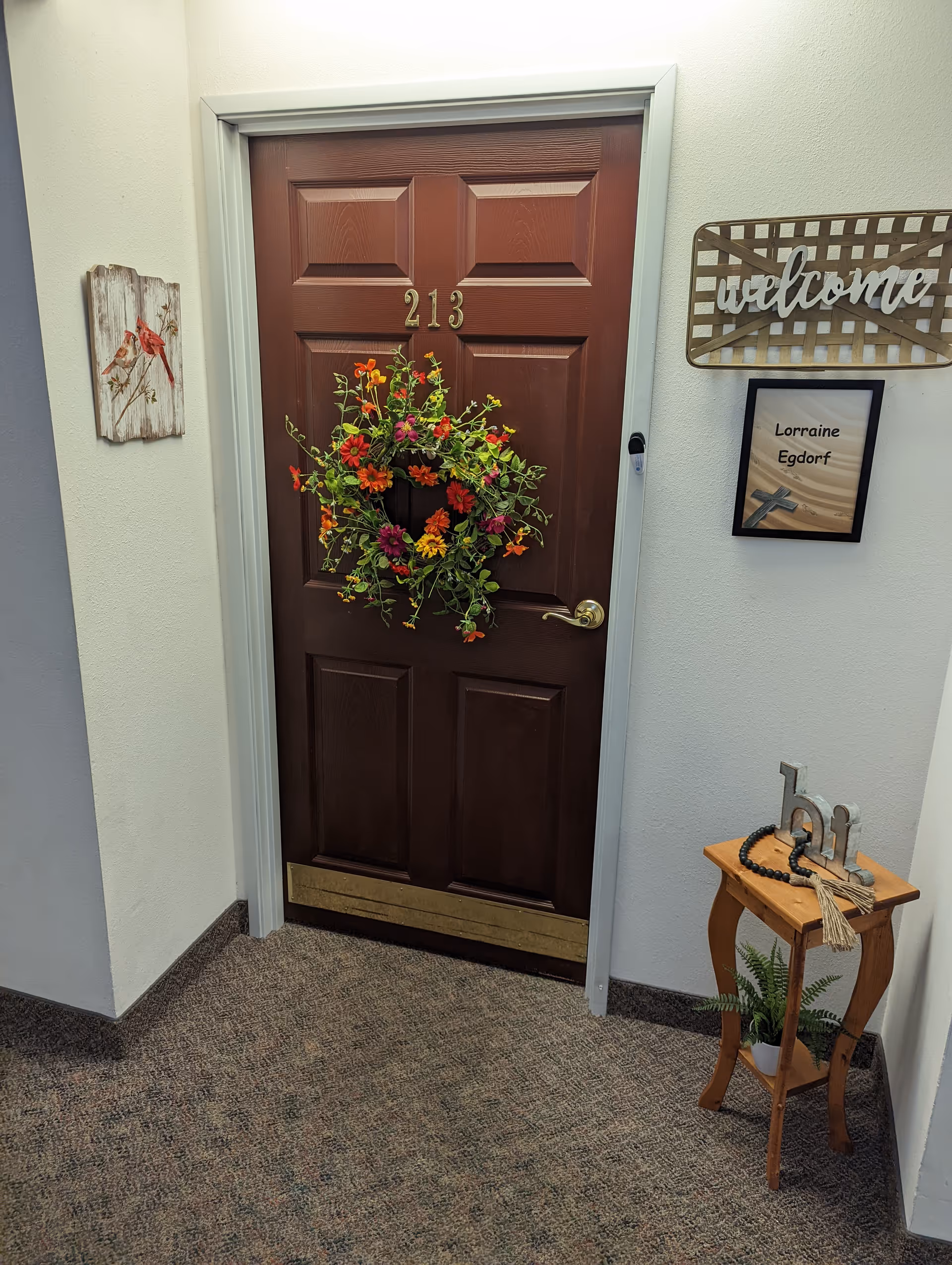 A brown door numbered 213 decorated with a colorful floral wreath. To the right of the door is a small wooden table with a plant and decorative items, and above it a wall sign that says 'welcome' and a framed name plaque reading 'Lorraine Egdorf'. On the left wall, there is a small artwork featuring birds.