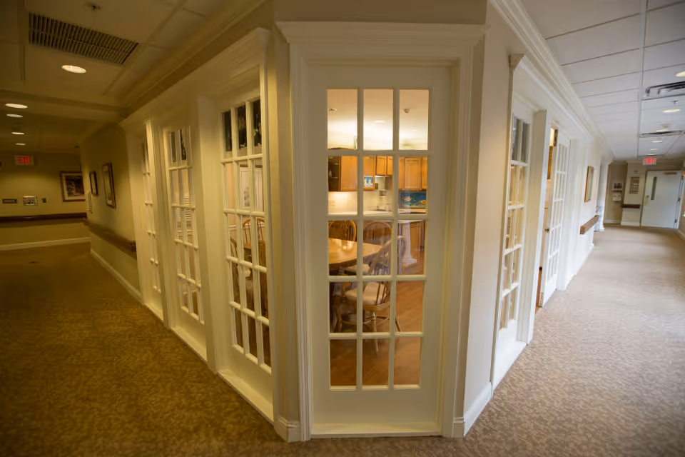 Interior hallway of a senior living facility with beige carpet and walls, featuring multiple white-framed glass doors. Through the glass doors, a dining area with a wooden table and chairs is visible, along with a kitchen area with wooden cabinets and a countertop.