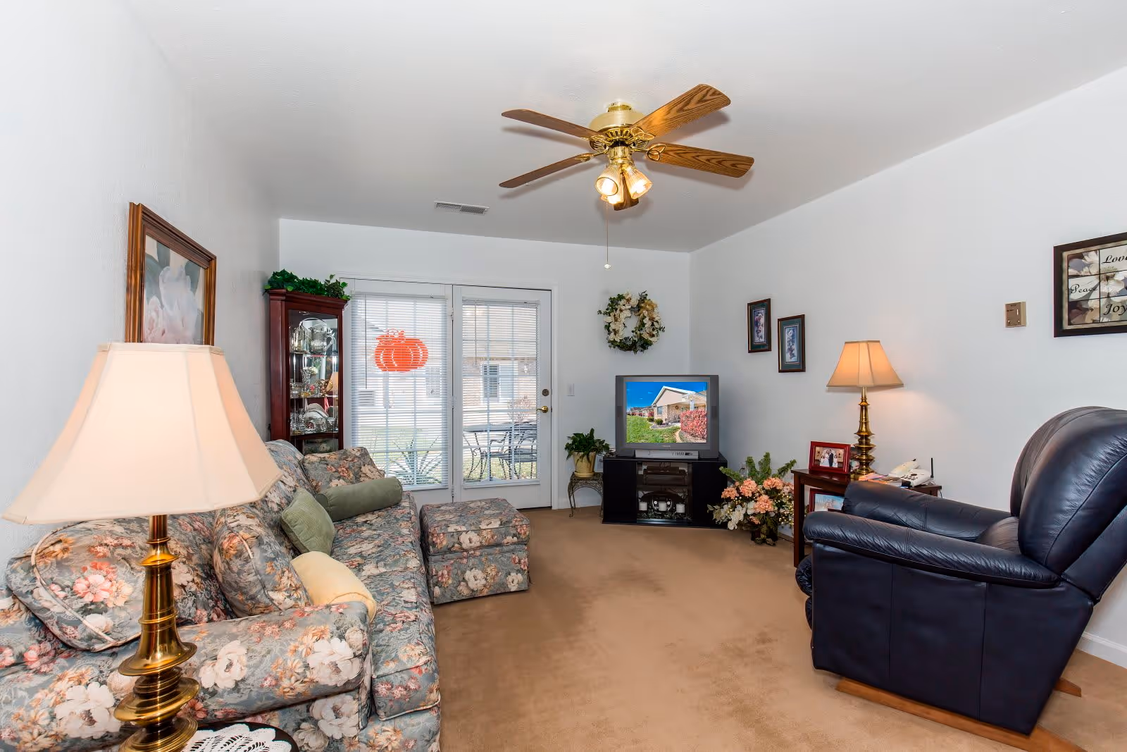 A cozy living room with a floral patterned sofa and matching ottoman, a dark leather armchair, two brass table lamps with beige lampshades, a ceiling fan with lights, a TV on a black stand displaying a house image, a glass display cabinet, and a glass door leading outside with a red pumpkin decoration on the window.