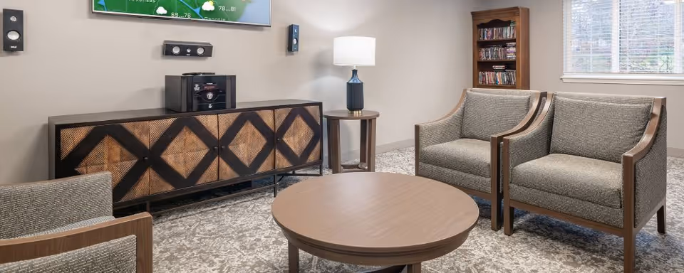 A cozy living room area with two gray upholstered armchairs, a round wooden coffee table, a wooden side table with a lamp, a wooden cabinet with a geometric pattern, a mounted TV, and a bookshelf filled with DVDs next to a window with blinds.