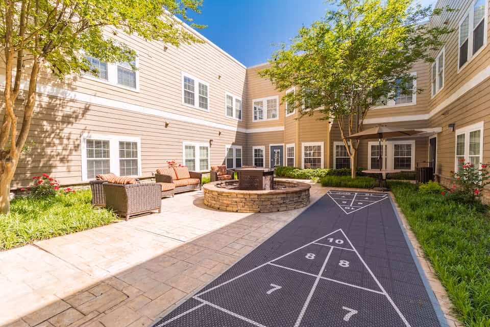 Outdoor courtyard area of a senior living facility with beige siding buildings surrounding it. The courtyard features a circular stone fire pit in the center, wicker seating with cushions, a shuffleboard court, green plants, and trees under a clear blue sky.