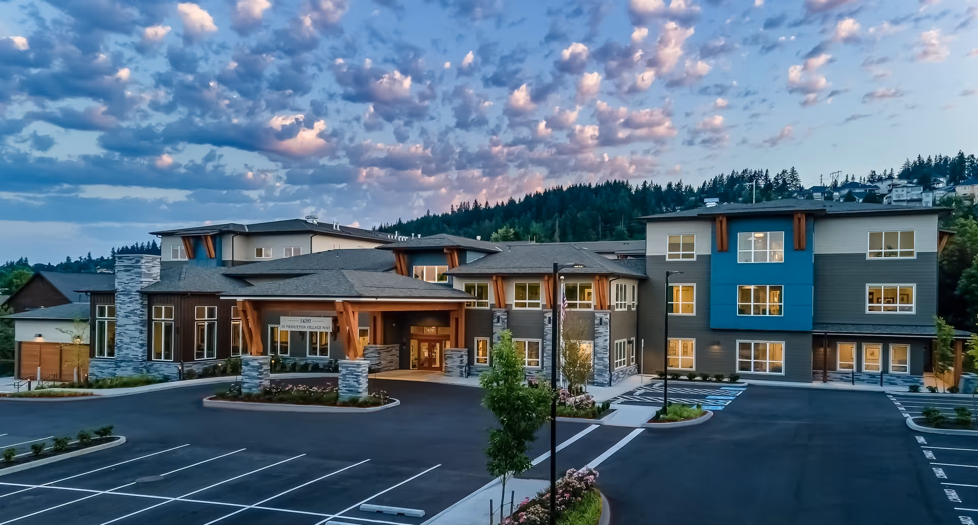 Exterior view of a modern assisted living and memory care facility building during dusk, with a large parking lot in front and a scenic backdrop of trees and hills under a partly cloudy sky.