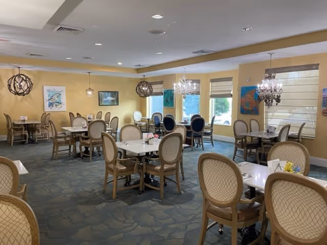A dining room with multiple tables and chairs arranged neatly. The room has yellow walls adorned with colorful paintings and large windows with beige blinds allowing natural light to enter. Decorative light fixtures hang from the ceiling, and the floor is covered with a patterned carpet.