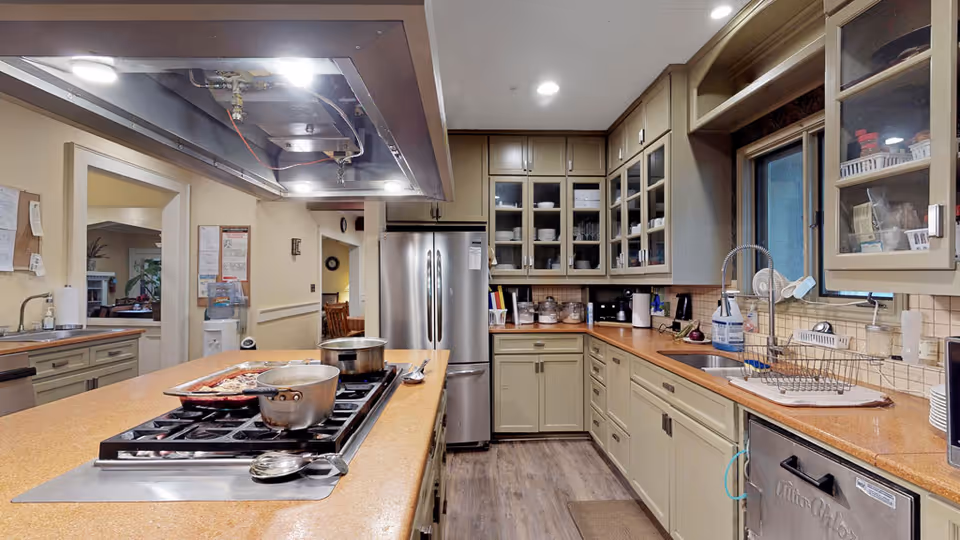 Communal kitchen with a long island cooktop holding pots, stainless steel refrigerator, glass-front cabinets, and a sink with a dish rack.
