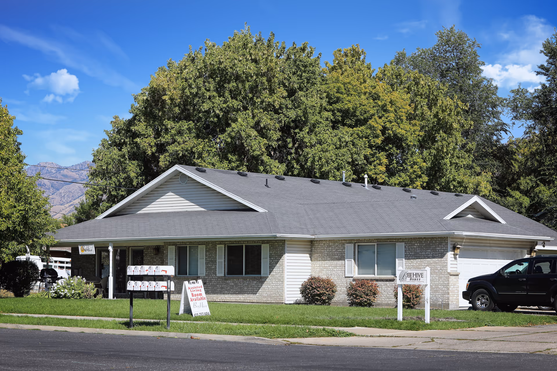 Single-story residential building with a gray roof and light-colored brick and siding exterior, surrounded by green grass and trees under a blue sky. There are multiple mailboxes and signs in front of the building, one of which reads 'Beehive Homes'. A black SUV is parked in the driveway.