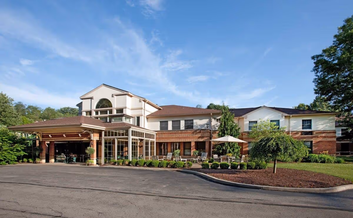 Exterior view of The Pines of Mount Lebanon facility showing a two-story building with a covered entrance, large windows, and outdoor seating area with chairs and an umbrella, surrounded by greenery and trees under a blue sky.