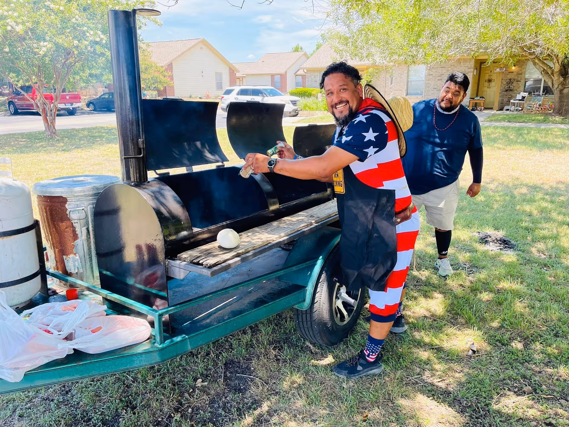 Two men are outdoors next to a large black barbecue smoker grill. One man, wearing a patriotic outfit with stars and stripes and a straw hat, is smiling and tending the grill. The other man stands behind him on the grass, looking towards the camera. There are houses, cars, and trees in the background.