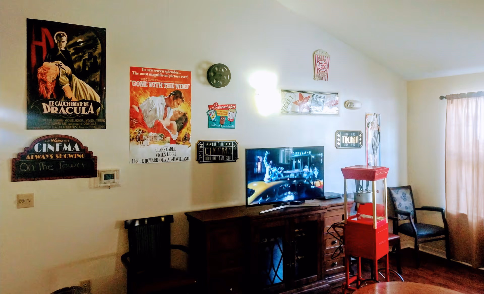 A cozy room with movie-themed decorations on the wall including posters for Dracula and Gone With The Wind, cinema signs, and a popcorn machine. There is a TV on a wooden cabinet showing a scene from a movie, two chairs, and a window with curtains letting in natural light.