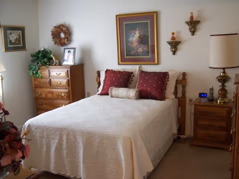A cozy bedroom with a neatly made bed covered in a white quilt and adorned with two red pillows and a cylindrical bolster pillow. On the wall above the bed hangs a framed floral painting. To the right of the bed is a wooden nightstand with a lamp, an alarm clock, and decorative items. To the left is a wooden dresser with a plant, a framed photo, and a wreath hanging above it. The room has a warm and inviting atmosphere.