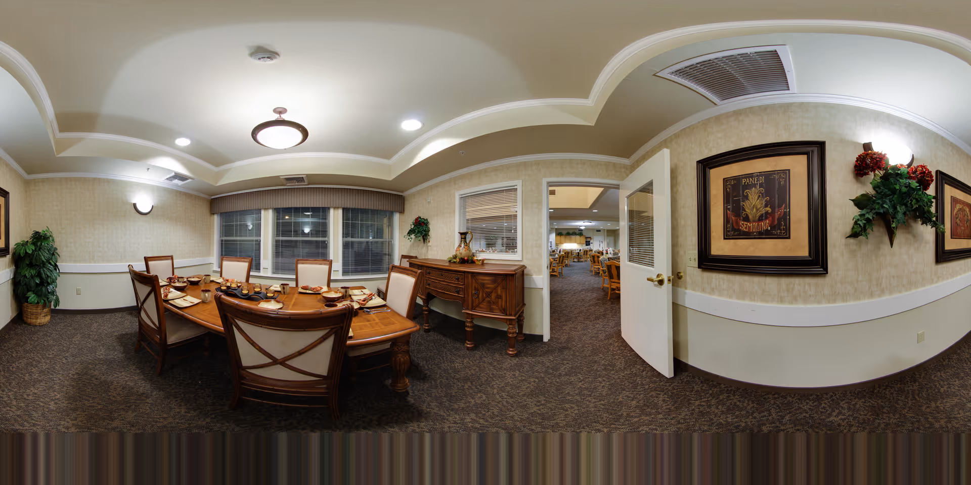 A cozy dining room in a senior living facility with a wooden dining table set for six people. The table is adorned with plates, cups, and a centerpiece. The room features beige walls with framed artwork, a potted plant in the corner, and a sideboard with decorative items. A door is open, revealing another dining area with multiple tables and chairs.