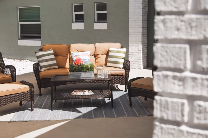 Outdoor patio area with cushioned wicker seating including a loveseat and chairs arranged around a wicker coffee table on a blue striped rug. The loveseat has three pillows, two with green and white stripes and one with a red chair design. There are two glasses of water with lemon slices on the table. The background shows a green wall with windows and a white brick column.