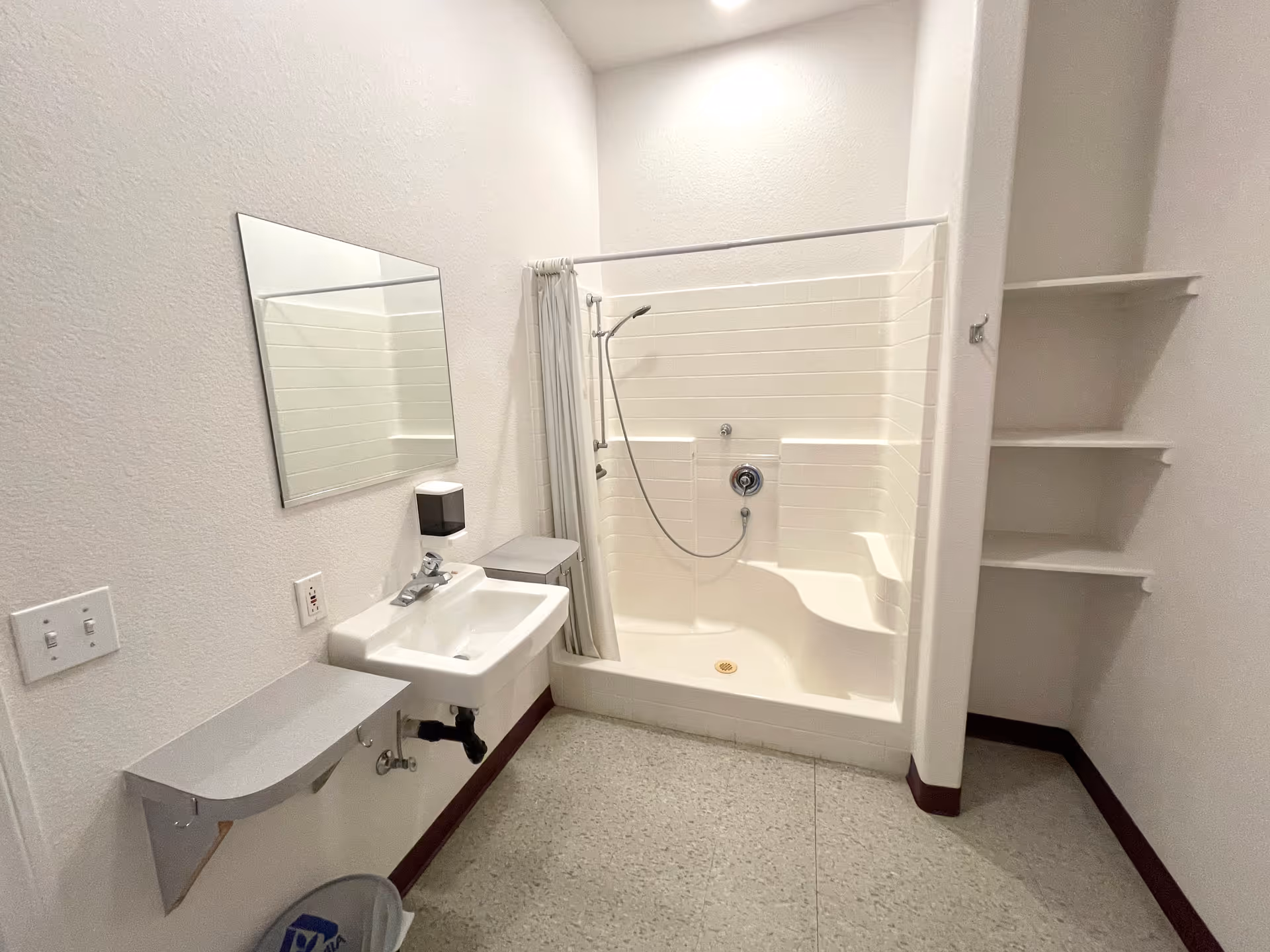 A clean, white bathroom featuring a walk-in shower with a built-in seat and handheld showerhead, a wall-mounted sink with a mirror above it, a soap dispenser, and open shelving on the right side. The floor is light-colored with a speckled pattern.
