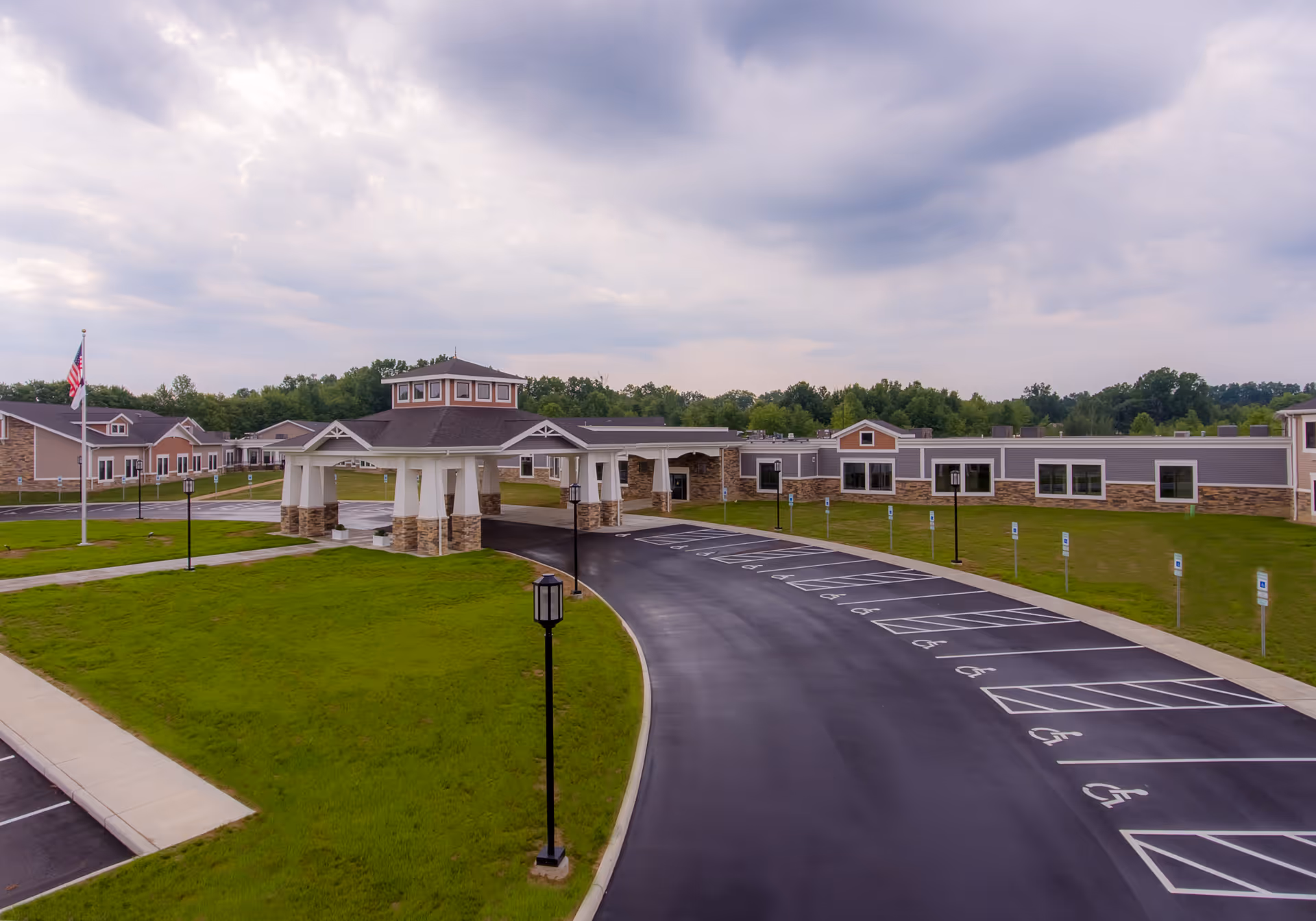 Exterior view of Shepherd of The Valley - Liberty facility showing a large covered entrance with stone pillars, a curved driveway with multiple handicap parking spaces, green lawns, and an American flag on a flagpole under a cloudy sky.