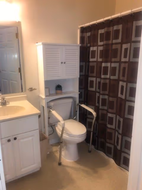 Bathroom with a white toilet equipped with safety rails, a white cabinet above the toilet, a sink with a countertop and cabinet below, a mirror above the sink, and a shower with a brown and beige geometric patterned curtain.