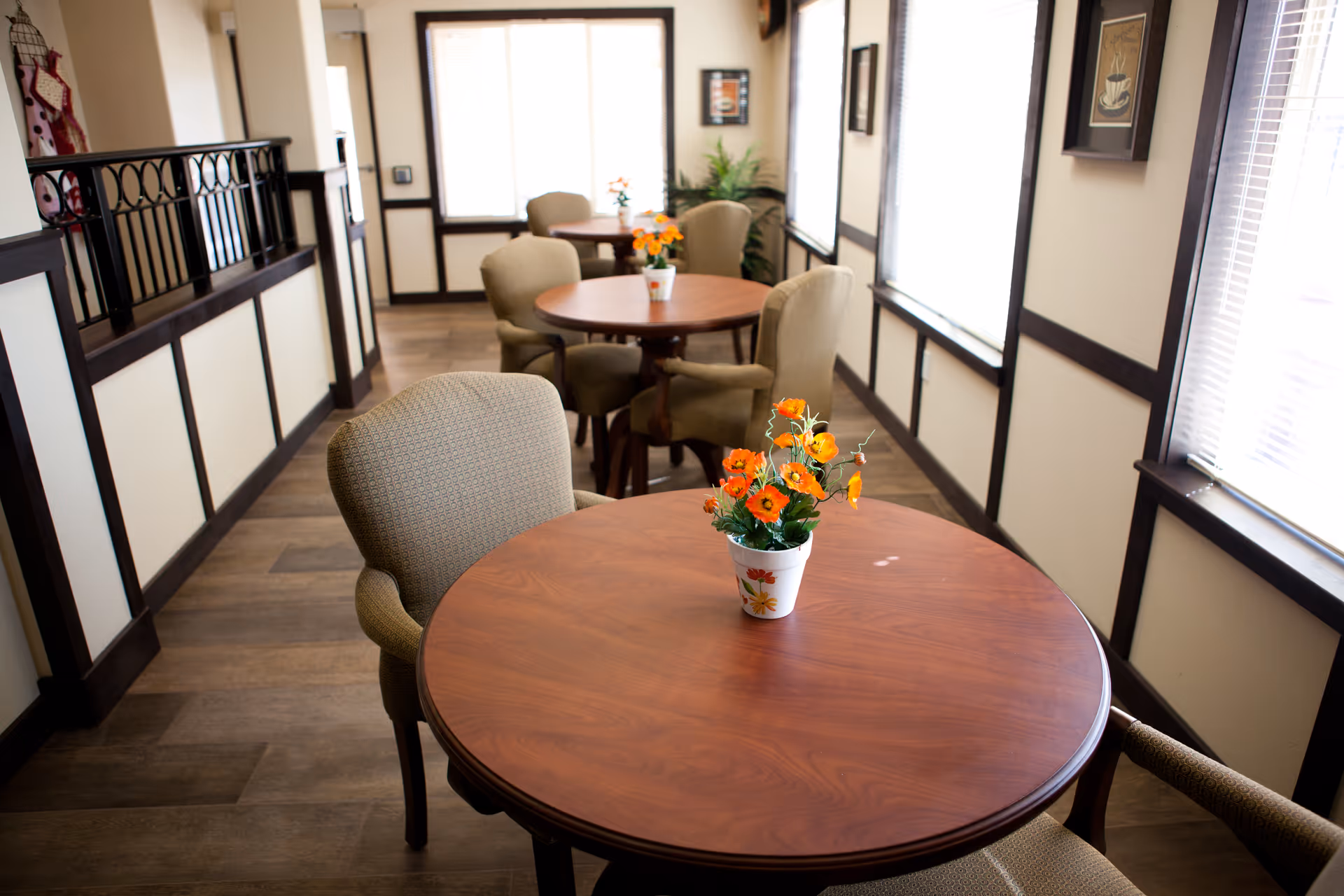 Round wooden tables with upholstered chairs and small potted flower centerpieces in a bright communal dining area.