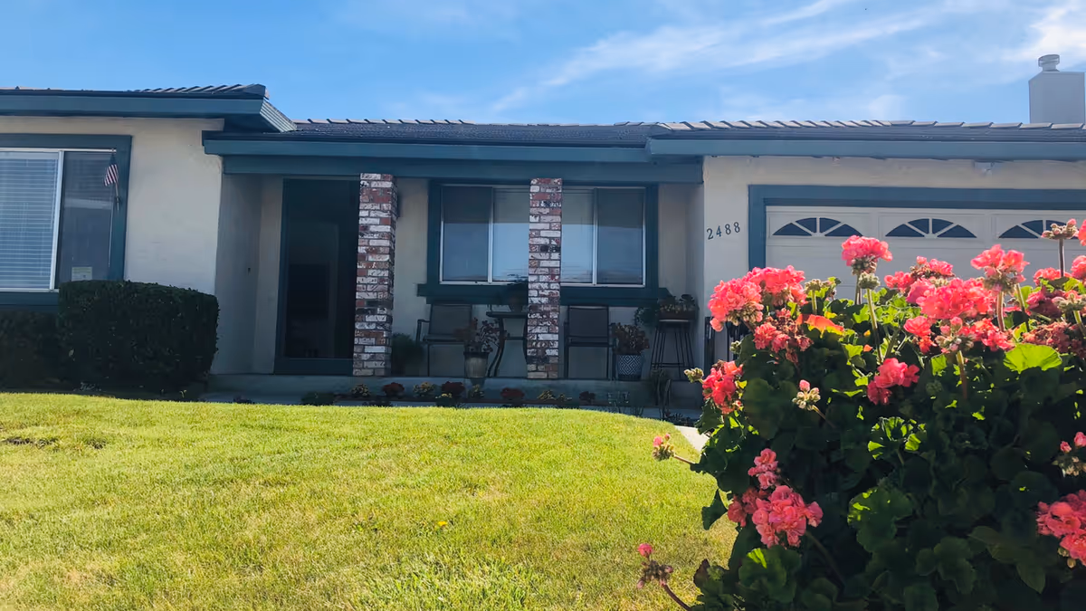 Front exterior view of a single-story residential care facility with a green lawn and blooming pink flowers in the foreground. The building has a covered porch with two brick columns, a window, and a garage door. The address number 2488 is visible near the garage.