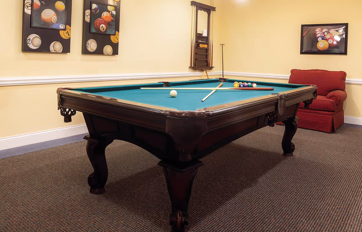 A billiards room with a pool table, cues and balls, framed pool artwork on the walls, and a red armchair.