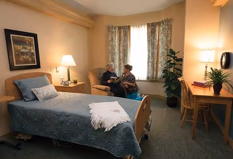 A cozy bedroom in a senior living facility with a single bed covered in a blue quilt and a white throw blanket. Two elderly women are sitting and talking near a window with floral curtains. The room also features a wooden desk with a chair, a potted plant, a table lamp, and a framed picture on the wall.