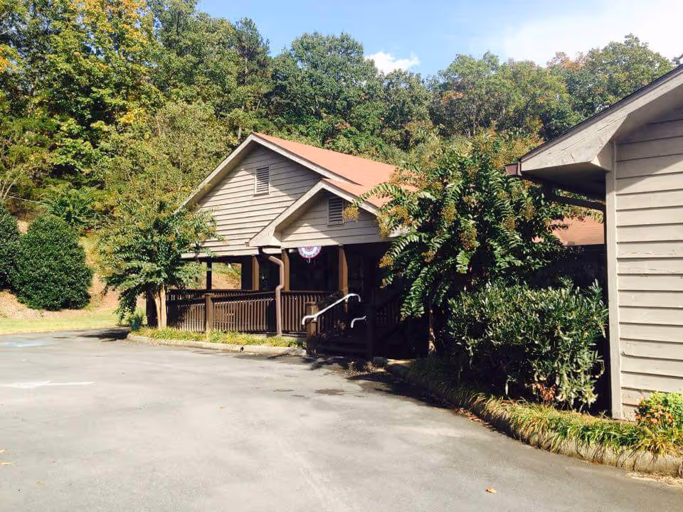 Front of a single-story wood-clad building with a covered porch, surrounded by trees and a paved driveway.