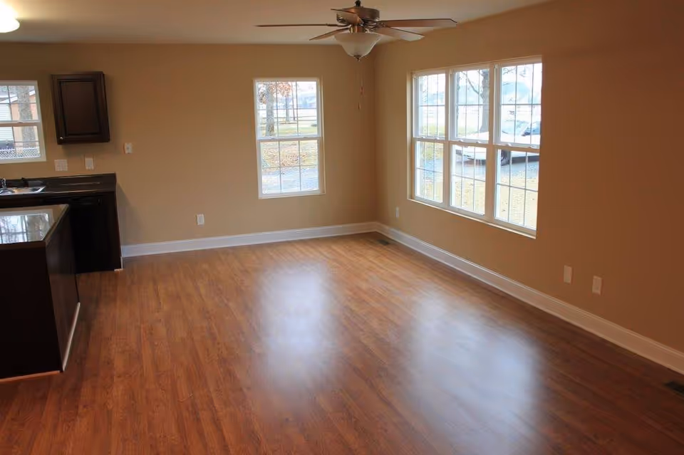 Empty room with wooden flooring, beige walls, and multiple windows letting in natural light. There is a ceiling fan with a light fixture in the center of the ceiling. On the left side, part of a kitchen area with dark cabinets and a countertop is visible.