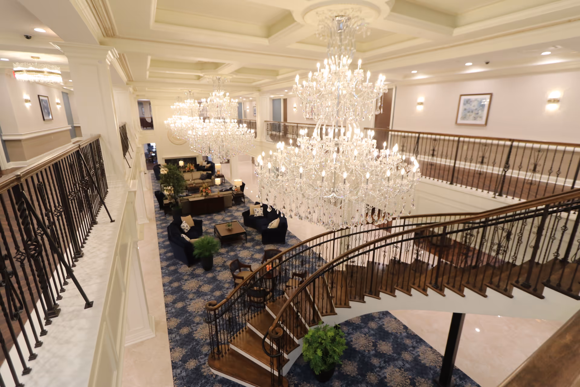 View from the upper level of an elegant senior living facility lobby featuring a curved wooden staircase with wrought iron railings, large crystal chandeliers, blue patterned carpet, and seating areas with navy blue chairs and sofas arranged around coffee tables. The walls are decorated with framed artwork and sconces, and there are several potted plants adding greenery to the space.