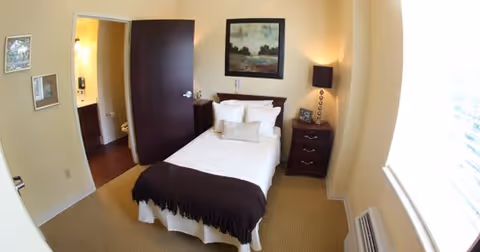 A small bedroom with a single bed covered with white linens and a dark brown throw blanket. There are multiple pillows on the bed. Next to the bed is a dark wooden nightstand with a lamp and decorative items. A framed painting hangs above the bed. The room has beige walls and carpet, a large window with blinds, and an open door leading to a bathroom.
