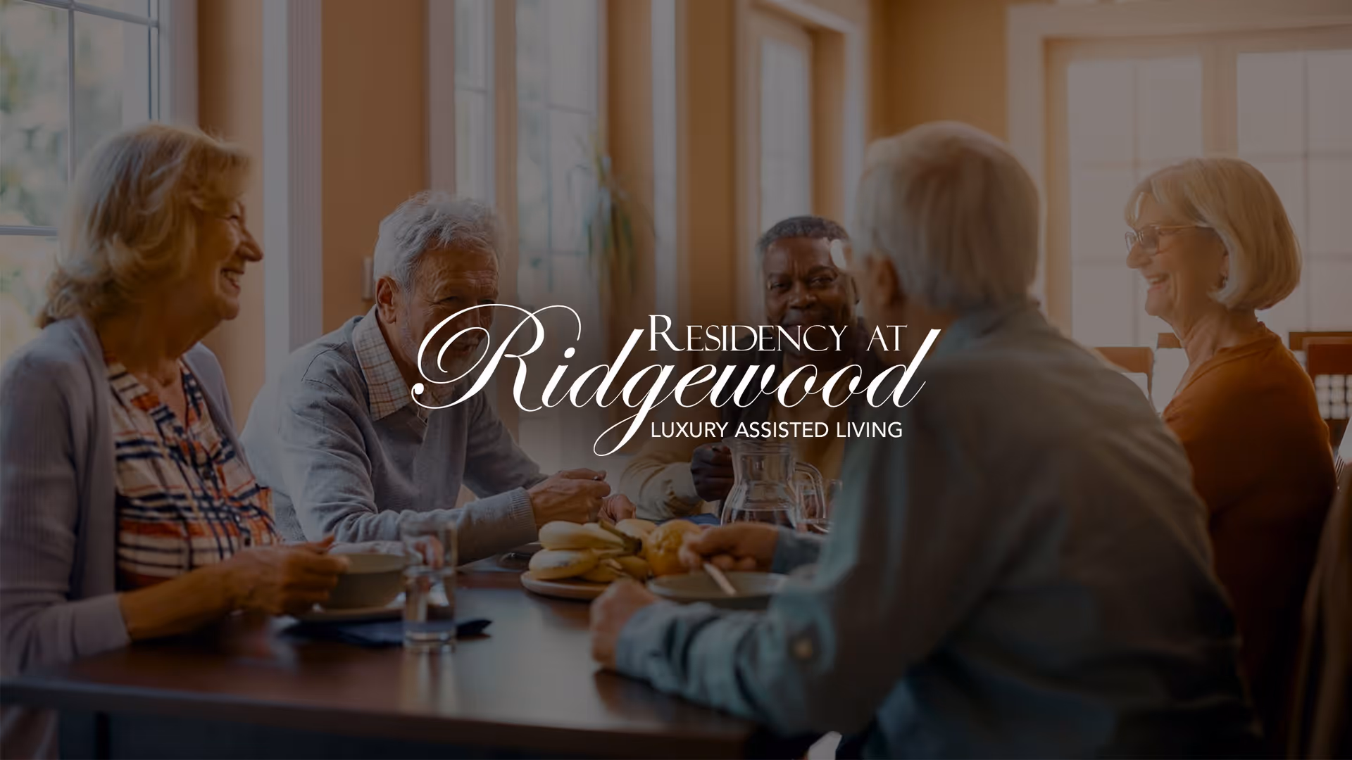 A group of older adults sitting around a dining table, smiling and chatting in a bright room.